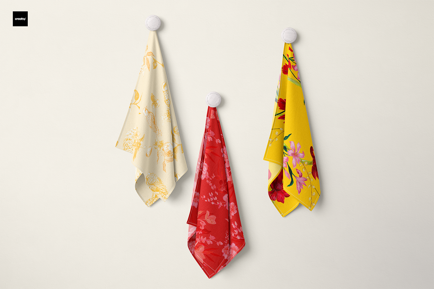 Three tea towels hanging on hooks: one with a bold red floral design, one featuring a whimsical citrus print with lemons and birds, and one with a vibrant tropical floral pattern. Each design is hand-painted by the artist, showcasing bold, bright, and joyful prints.